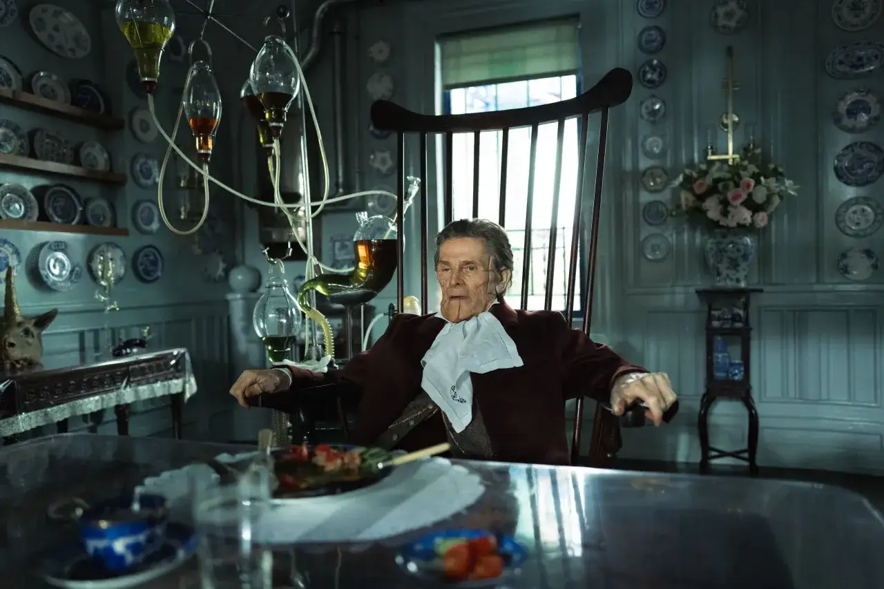 Willem Dafoe sitter i en gungstol i ett rum fyllt med antika tallrikar och laboratorieutrustning. Hans karaktär i filmen verkar vara en excentrisk vetenskapsman.