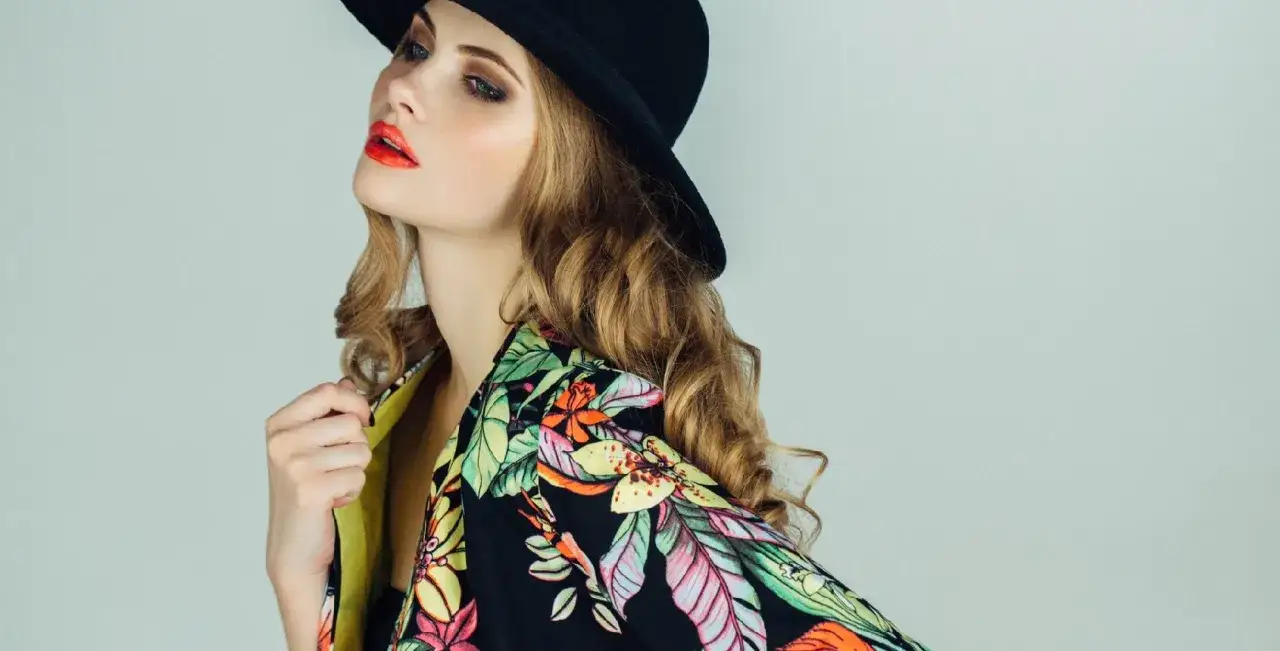 Mujer con sombrero negro y chaqueta estampada floral, luciendo un labial rojo vibrante. ¡Un look perfecto para tu próximo custo outlet!