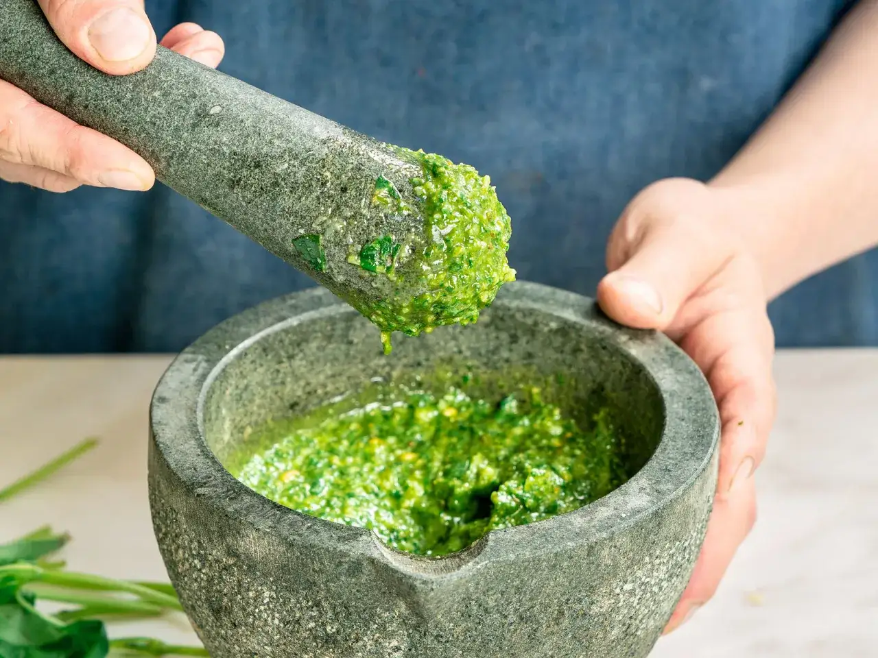 Frische Kräuter werden im Mörser zermahlen, um **Pesto selber machen** zu können. Ein Tropfen Öl löst sich vom Stößel.