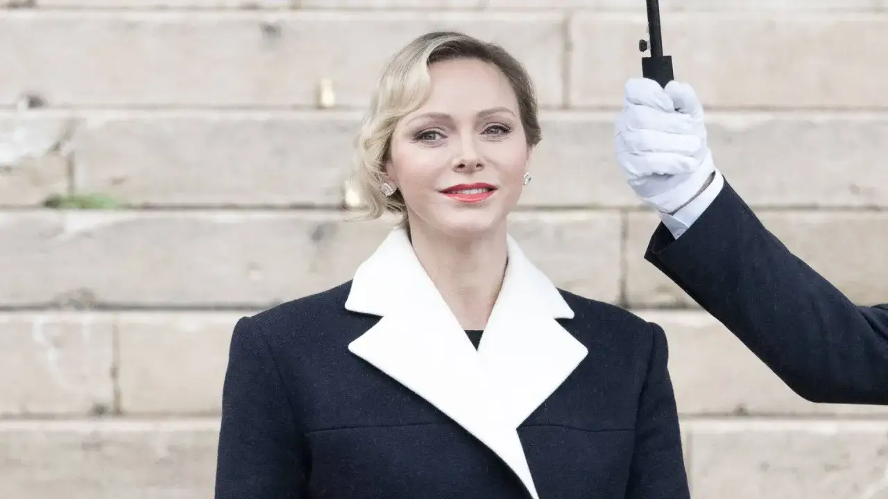 La princesa de Mónaco, elegante con abrigo oscuro y cuello blanco, sonríe mientras un asistente sostiene un paraguas.