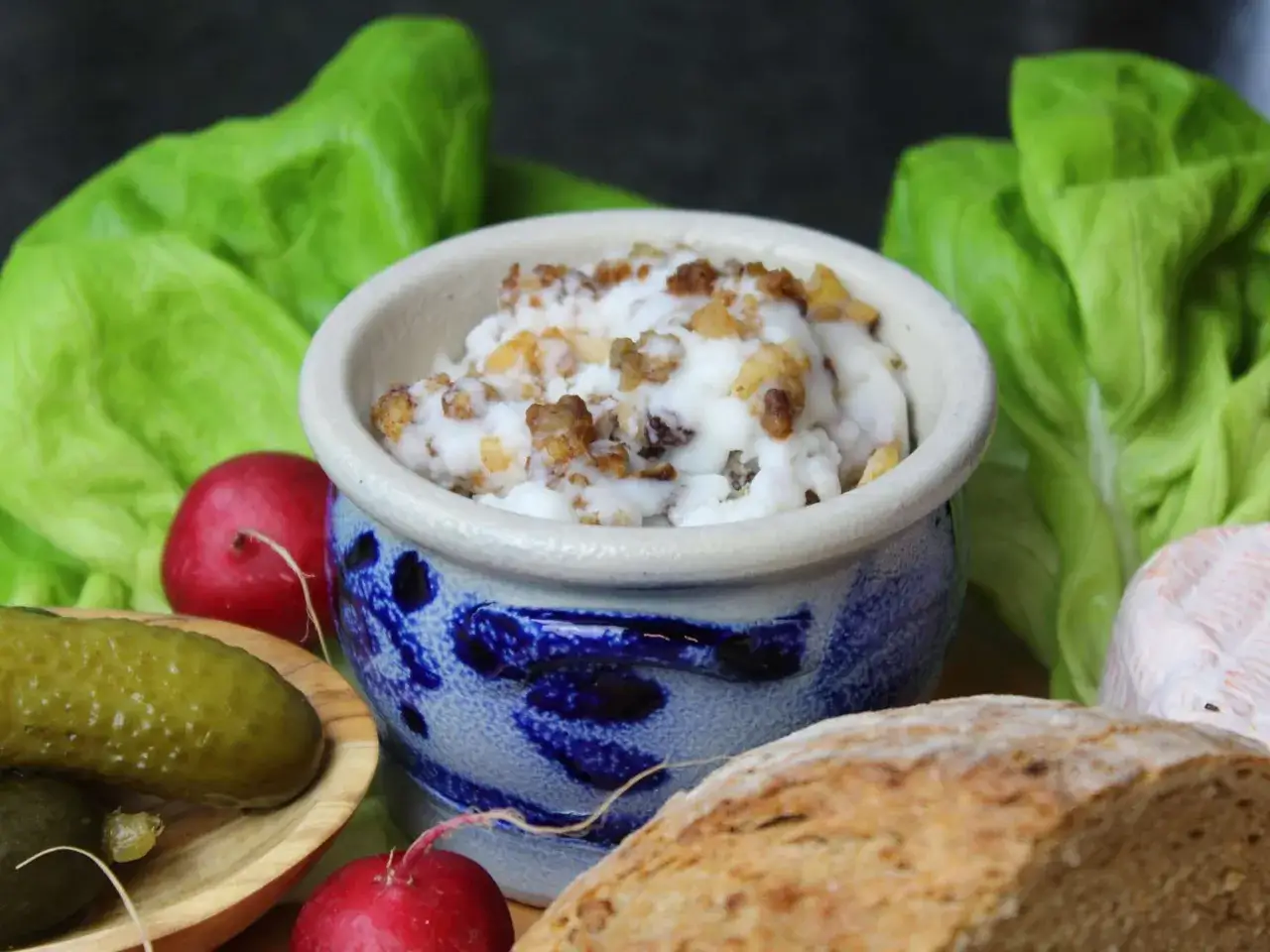 Schmalz mit knusprigen Grieben in einem blauen Steinguttopf, umgeben von Salat, Radieschen, Gewürzgurken und Brot. Perfekt, um die Flomen auszulassen!