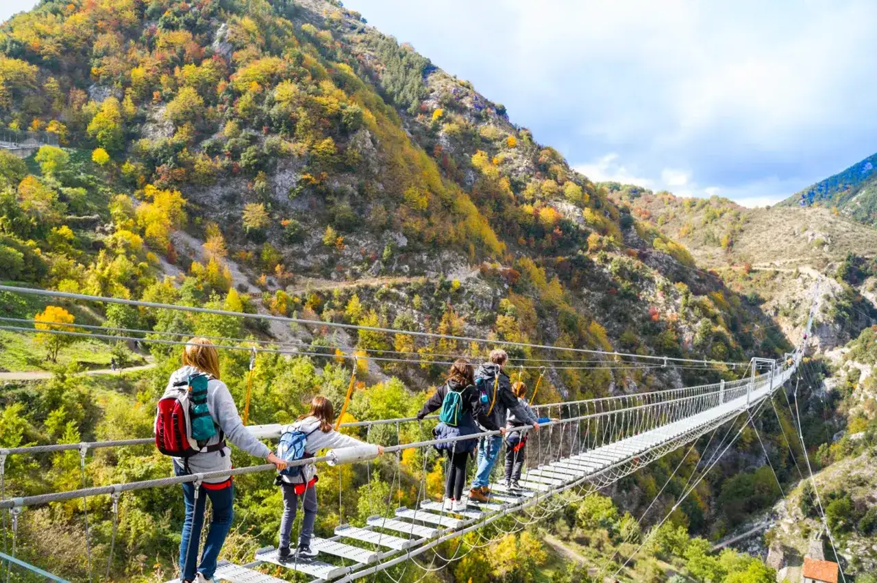 Famiglia attraversa un lungo ponte tibetano originale, circondato da un paesaggio montano autunnale dai colori vivaci.