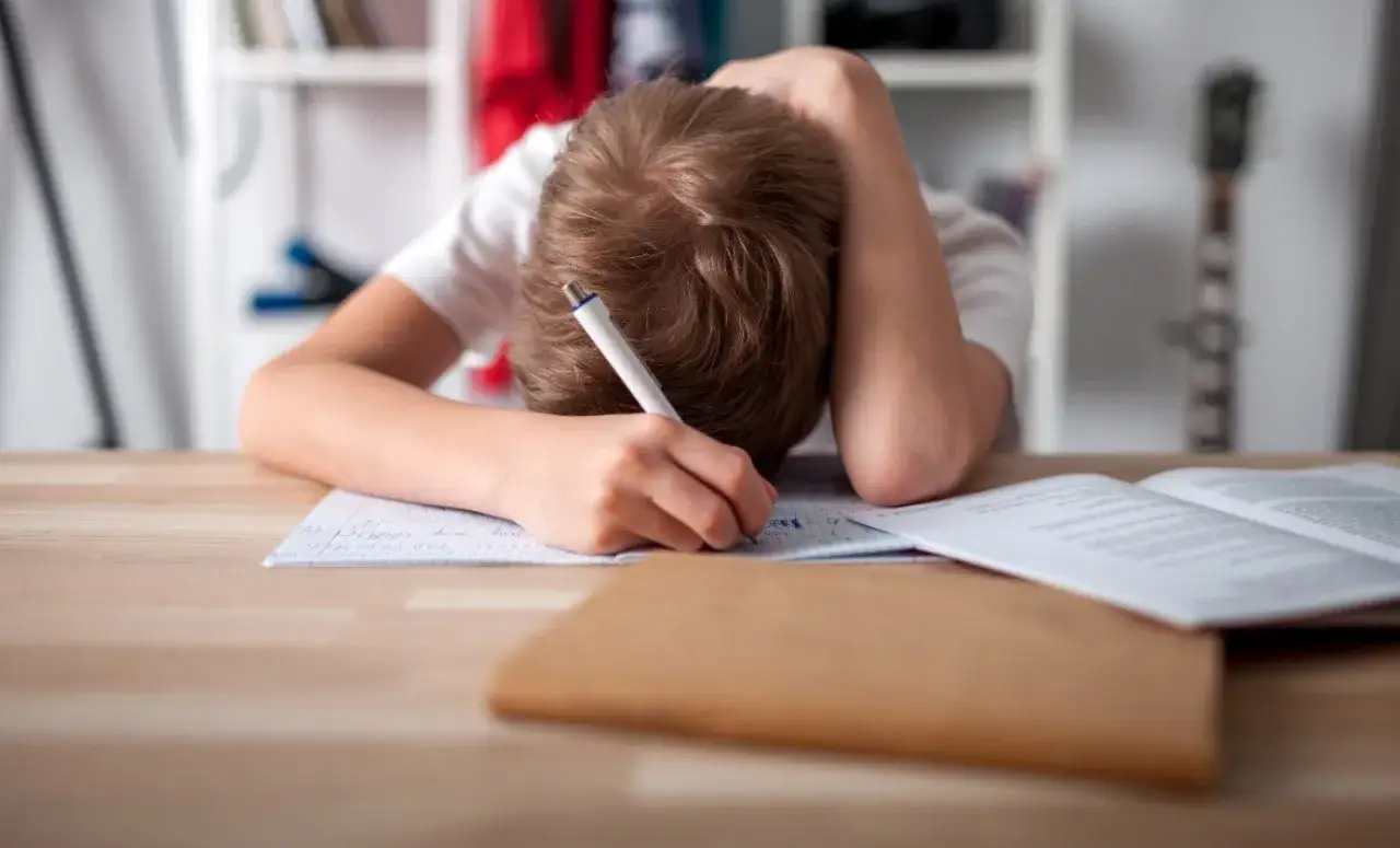 Chłopiec z ręką na głowie pisze w zeszycie, wyraźnie widać, że dziecko nie chce się uczyć. Psycholog mógłby pomóc.