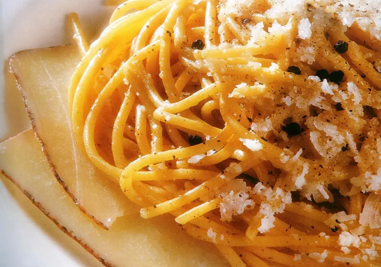 Pyszne spaghetti cacio e pepe z tartym serem i czarnym pieprzem, obok plastr&oacute;w sera.