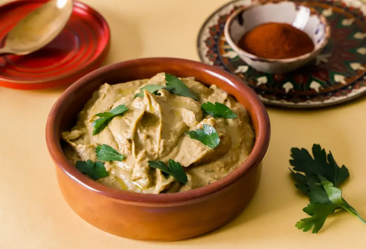 Un cremoso humus di lenticchie, guarnito con prezzemolo fresco, pronto per essere gustato. Accanto, una ciotolina con paprika e un cucchiaio su un piattino rosso.