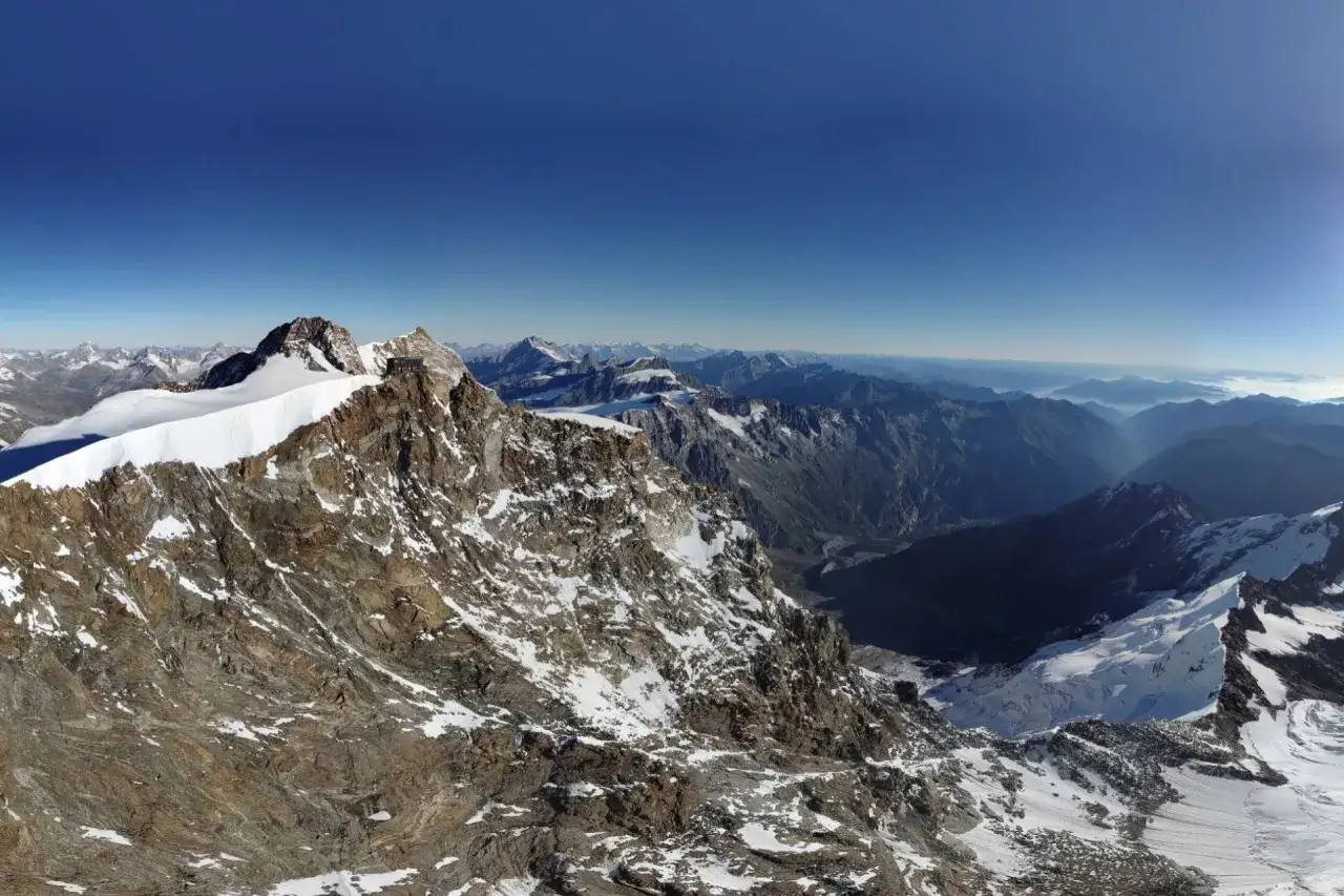 Majest&auml;tische Berglandschaft mit der Capanna Margherita auf einem Gipfel, umgeben von schneebedeckten Felsen und tiefen T&auml;lern unter blauem Himmel.