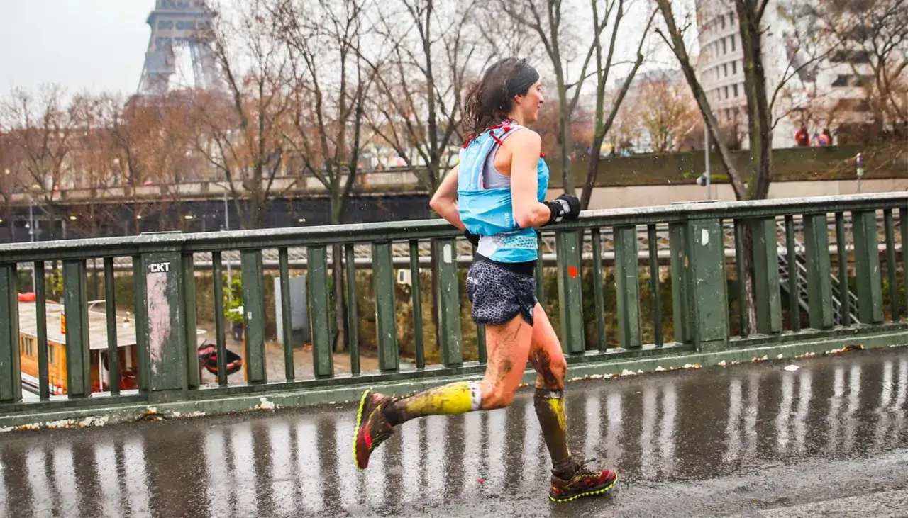 Une coureuse boueuse traverse un pont à Paris, la Tour Eiffel en arrière-plan. Elle court sous la pluie, ses jambes couvertes de terre.