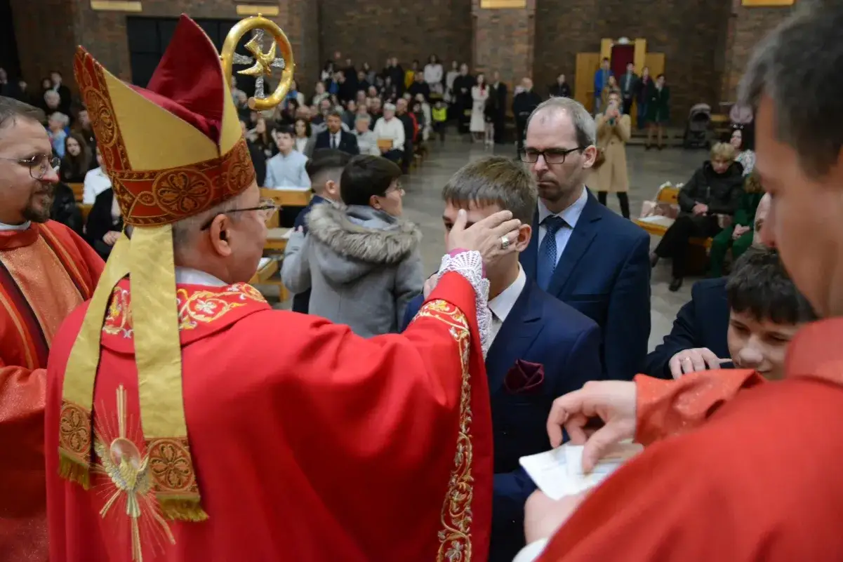Biskup udziela sakramentu bierzmowania chłopcu w kościele. W tle zgromadzeni wierni.