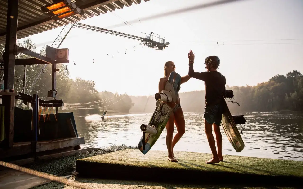 Zwei Wakeboarder geben sich am Langenfelder See einen High-Five. Im Hintergrund f&auml;hrt jemand &uuml;ber das Wasser.