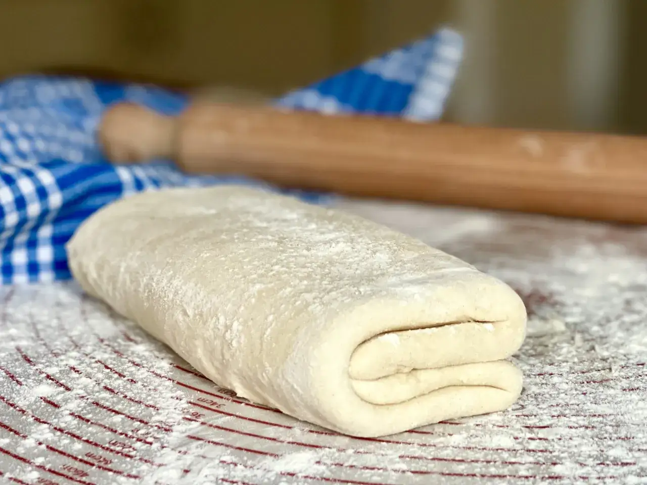 Impasto per pasta fatta in casa, piegato più volte, pronto per essere steso. Un mattarello e un canovaccio a quadretti blu sullo sfondo.