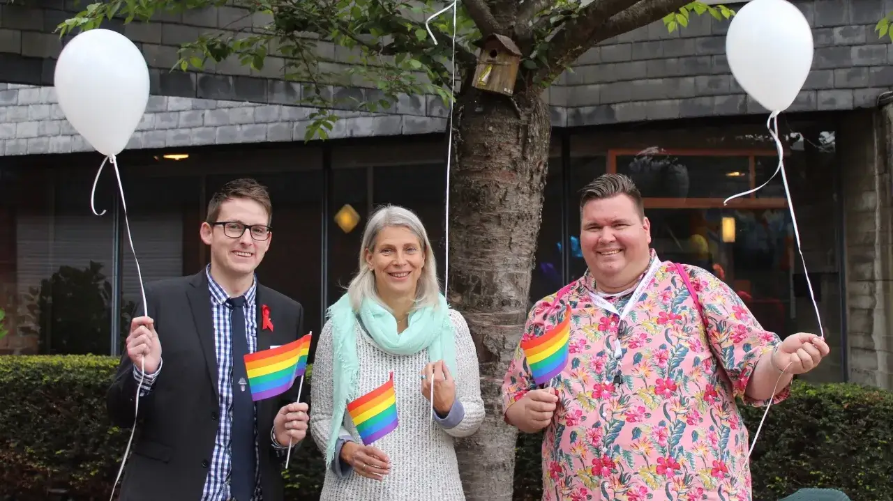 Drei Personen mit Regenbogenfahnen und wei&szlig;en Ballons vor einem Geb&auml;ude. Sie zeigen Unterst&uuml;tzung f&uuml;r die evangelische Kirche und die LGBT-Gemeinschaft.