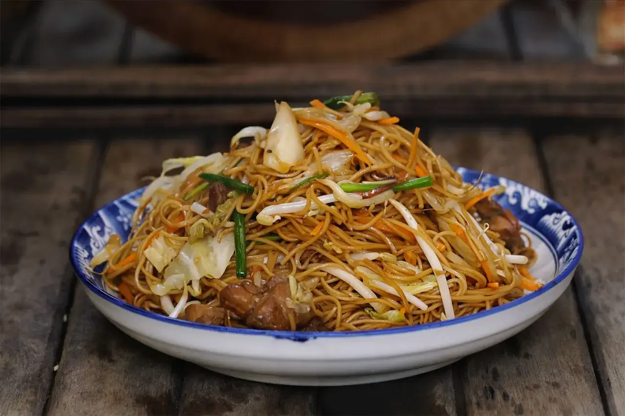 Un plato de chow mein con fideos dorados, trozos de carne, zanahoria, repollo y brotes de soja, servido en un cuenco azul y blanco.