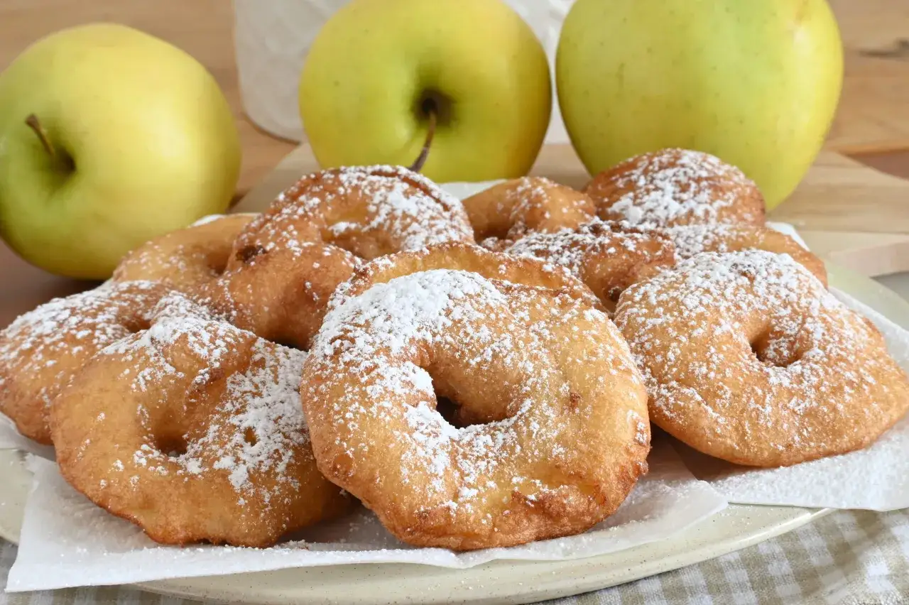 Un piatto di frittelle di mele dorate e spolverate di zucchero a velo, con mele verdi sullo sfondo.