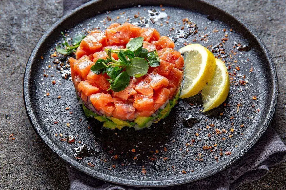 Tartare di salmone fresca e colorata, servita su un piatto nero con spicchi di limone e guarnita con erbe aromatiche.