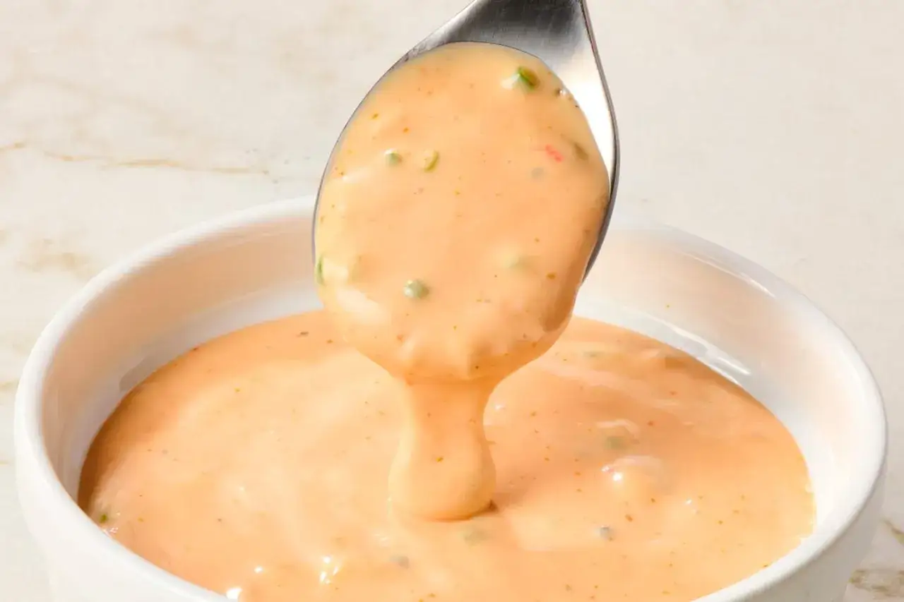 Ein Löffel cremig-orangenes Thousand Island Dressing tropft in eine weiße Schüssel. Kleine grüne und rote Stückchen sind sichtbar.