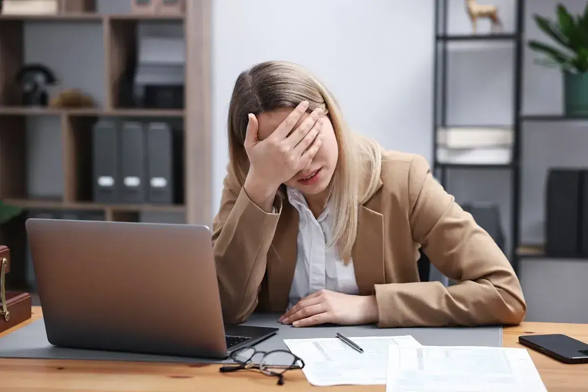 Zmęczona kobieta z ręką na czole przy laptopie. Czy na macierzyńskim można pracować? Trudne pytanie, gdy obowiązków jest tak wiele.