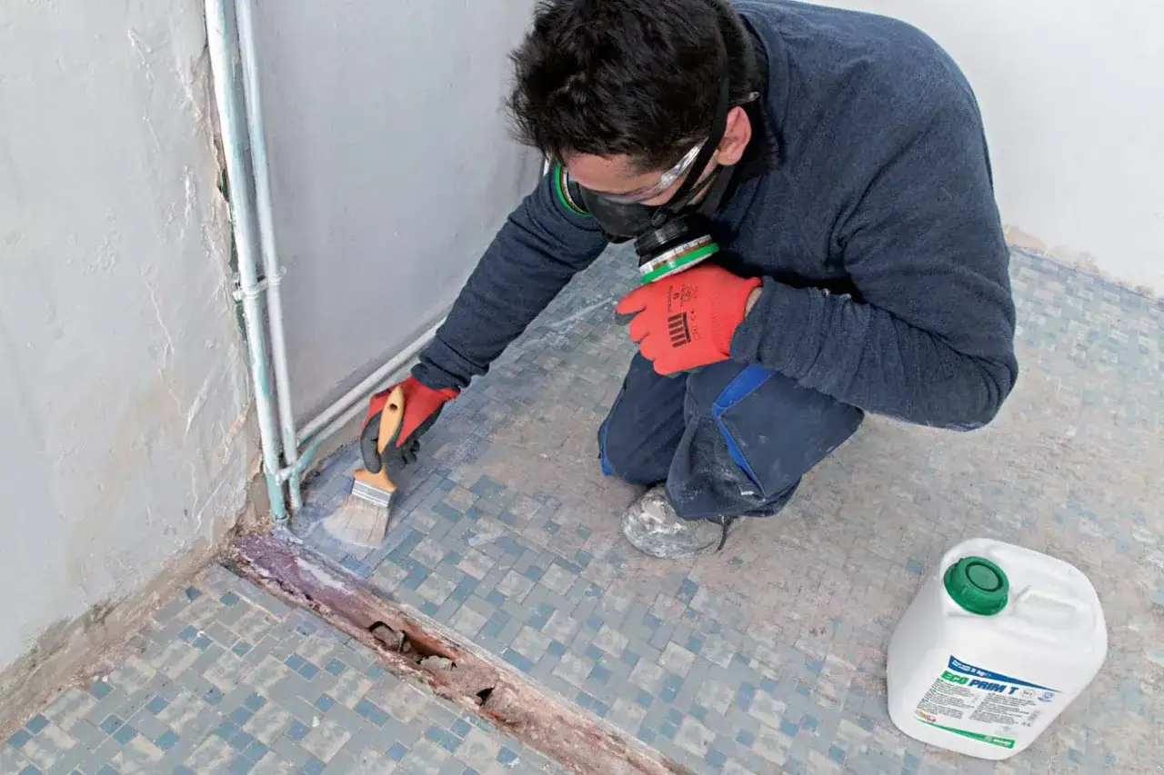 Un homme porte un masque et des gants pour appliquer un primaire avant un ragr&eacute;age sur carrelage.