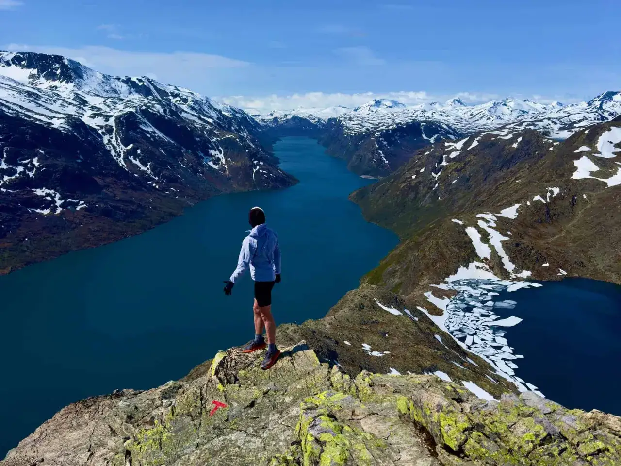 Vandrare p&aring; en klippavsats med utsikt &ouml;ver en djupbl&aring; sj&ouml; och sn&ouml;kl&auml;dda berg. En ikonisk vy fr&aring;n Besseggen, Norge.