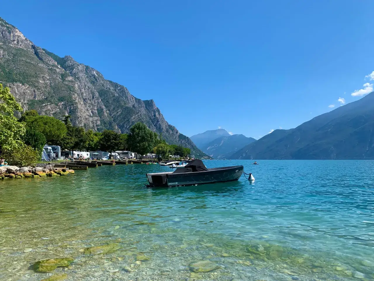 Spokojna tafla jeziora przy Punta San Vigilio, z łodzią na kotwicy i kempingiem w tle, otoczona górami.