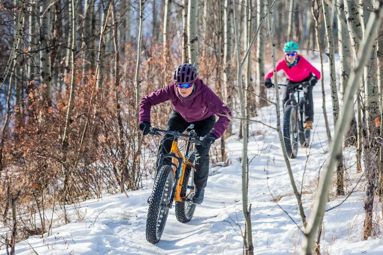 Two people enjoy a winter mtb ride through a snowy forest, their fat-tire bikes navigating the trail.