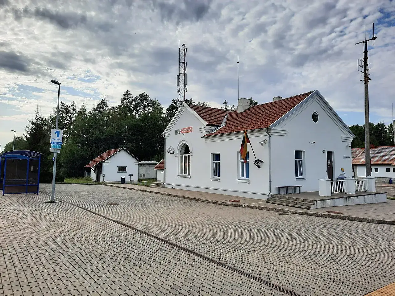 Stacja kolejowa Mockava z białym budynkiem, peronem i wiatą przystankową.
