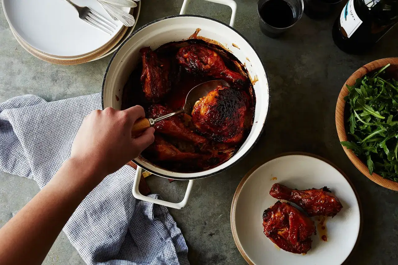 En hand serverar glaserad kyckling sambal oelek ur en gryta. Två kycklingdelar ligger på en tallrik bredvid.
