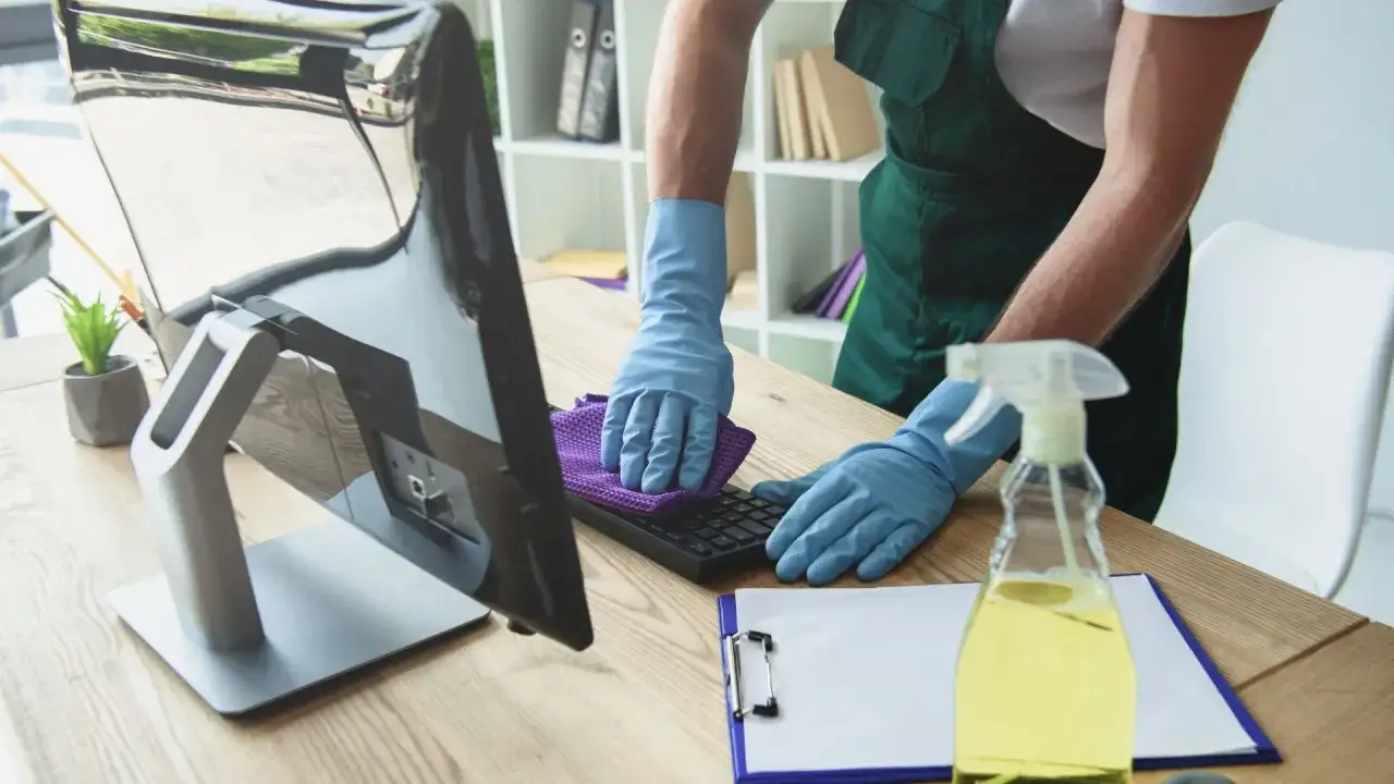 Un professionnel ganté utilise un chiffon violet pour nettoyer un clavier d'ordinateur, assurant un espace de travail hygiénique.