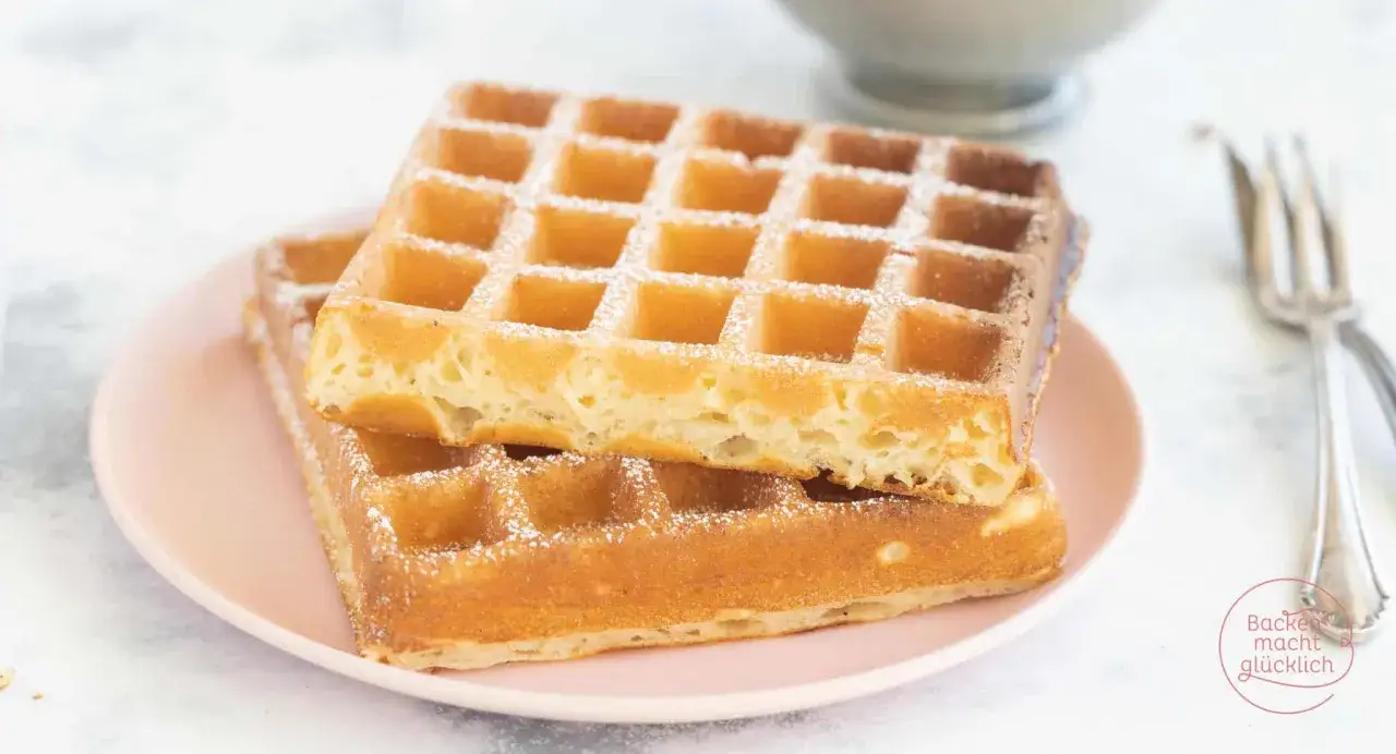 Zwei goldbraune belgische Waffeln, mit Puderzucker best&auml;ubt, auf einem rosa Teller gestapelt.