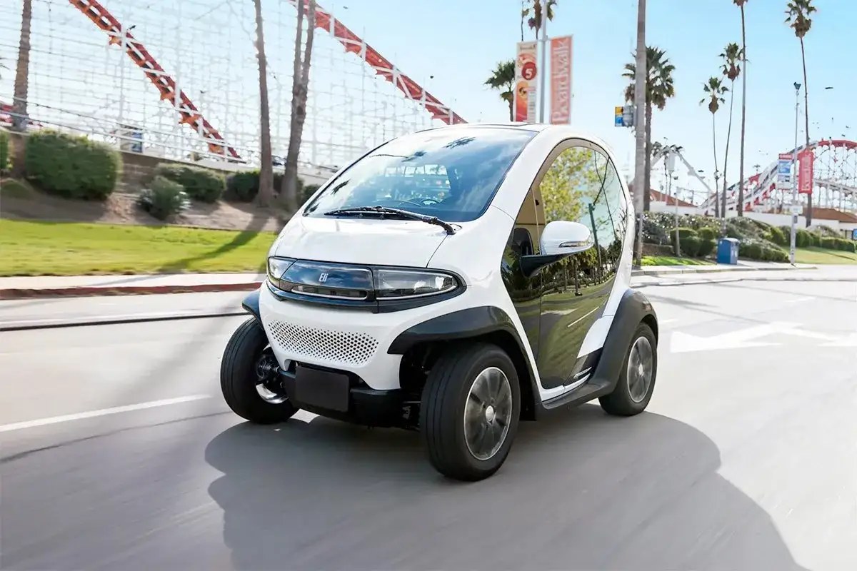 Biały, kompaktowy pojazd elektryczny Mitsubishi i-MiEV porusza się po drodze w słoneczny dzień, z widocznymi w tle palmami i atrakcjami parku rozrywki.