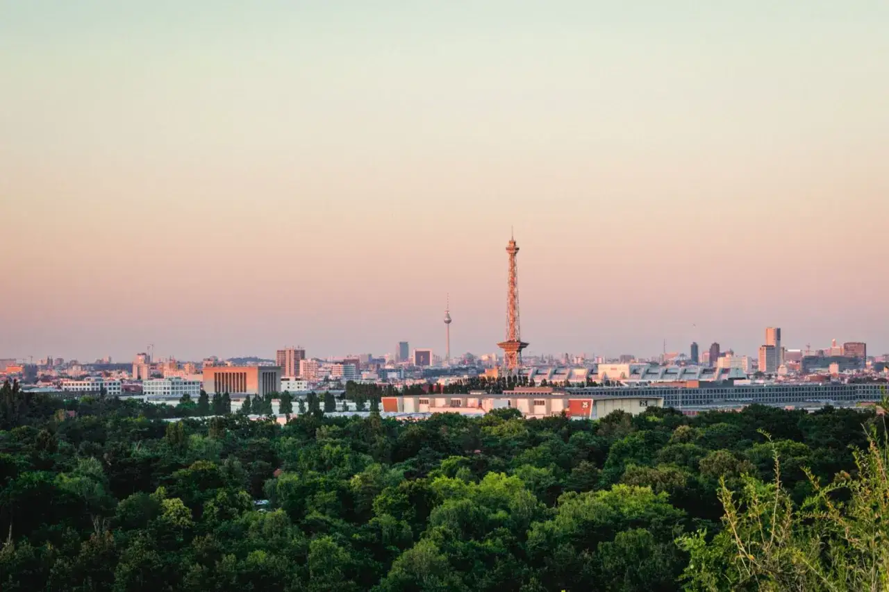 Blick vom Aussichtspunkt Drachenberg auf die Berliner Skyline bei Sonnenuntergang. Grüne Wälder im Vordergrund, Türme und Gebäude im Hintergrund.