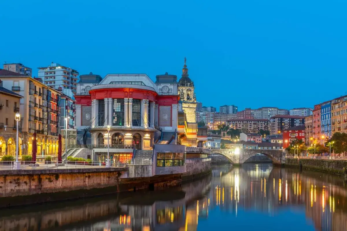 Wieczorny widok na rzekę Nervión w Bilbao, największym mieście Kraju Basków, z odbijającymi się w wodzie światłami budynków i mostu.