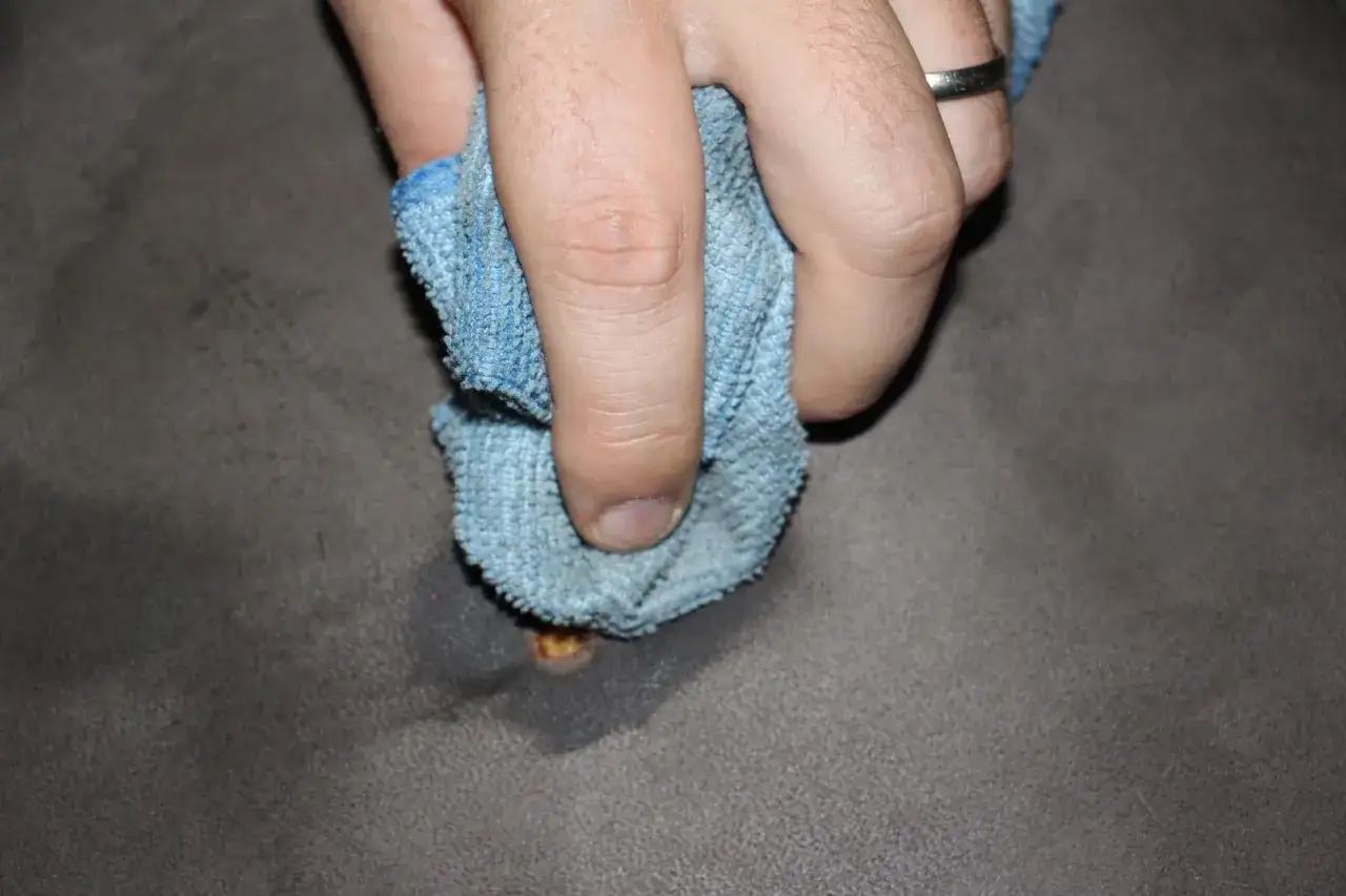 Mano con panno blu che pulisce una bruciatura da sigaretta su tessuto grigio.