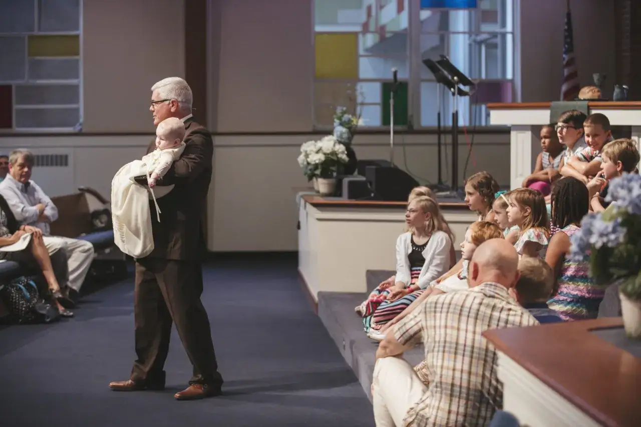 A pastor holds a baby in a baptismal gown, symbolizing the start of a spiritual journey. This event highlights 3 reasons why baptism is important: new beginnings, community welcome, and faith commitment.