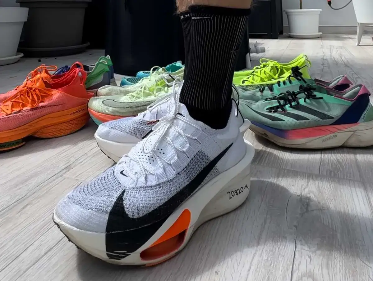 En person står i ett par vita löparskor, redo för hoka halvmaraton. Flera färgglada löparskor ligger utspridda bakom.