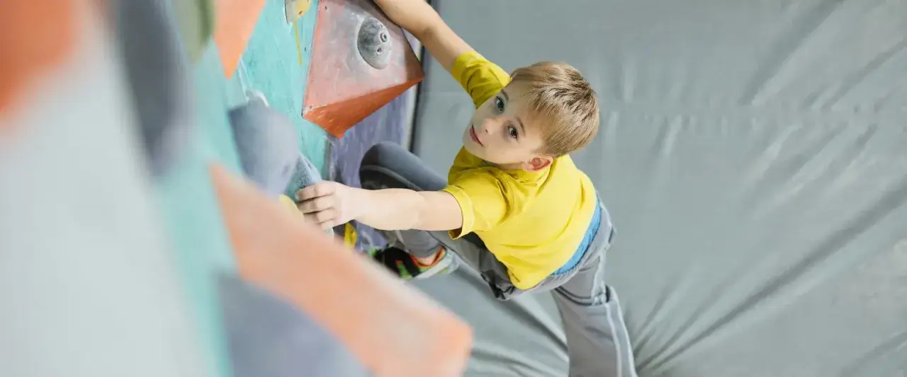 Bambino biondo in maglia gialla si arrampica su una parete colorata. Un piccolo scalatore, forse di 3 anni, si diverte con l'arrampicata.