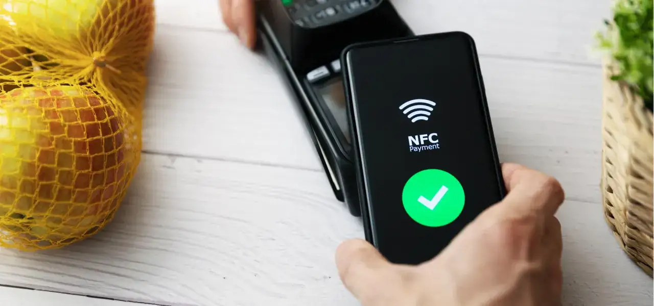 Płatność zbliżeniowa NFC telefonem przy kasie. Na ekranie telefonu widnieje zielony znaczek potwierdzenia.