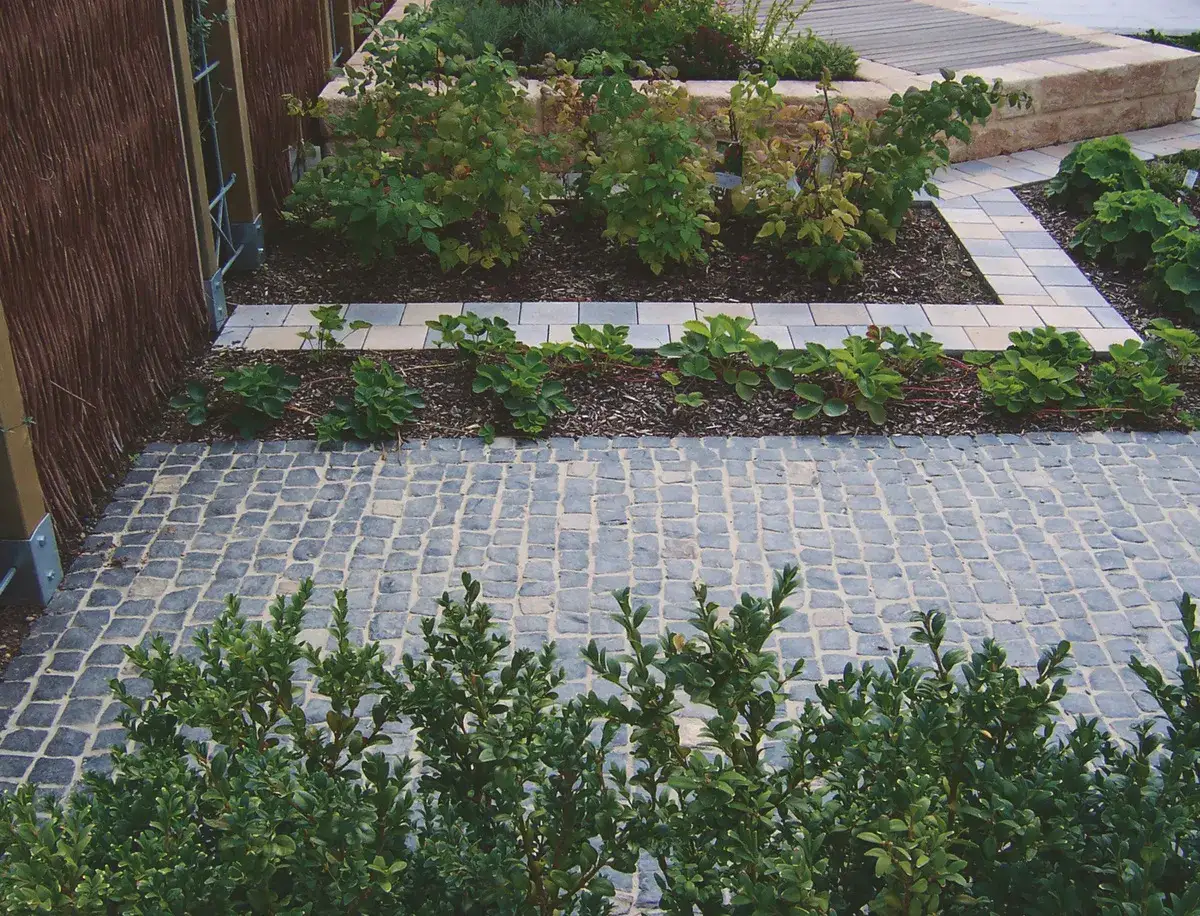 Ein Weg aus Kopfsteinpflaster, gefertigt aus dunklem Ergussgestein Basalt, führt durch einen Garten mit grünen Sträuchern und Beeten.