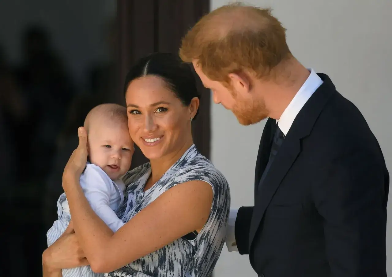 Meghan Markle z dzieckiem w ramionach, uśmiechnięta, obok księcia Harry'ego.
