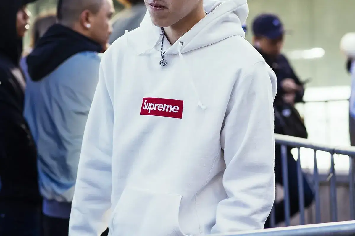 Młody mężczyzna w białej bluzie z kapturem i czerwonym logo Supreme. Supreme to marka znana z limitowanych kolekcji streetwear.