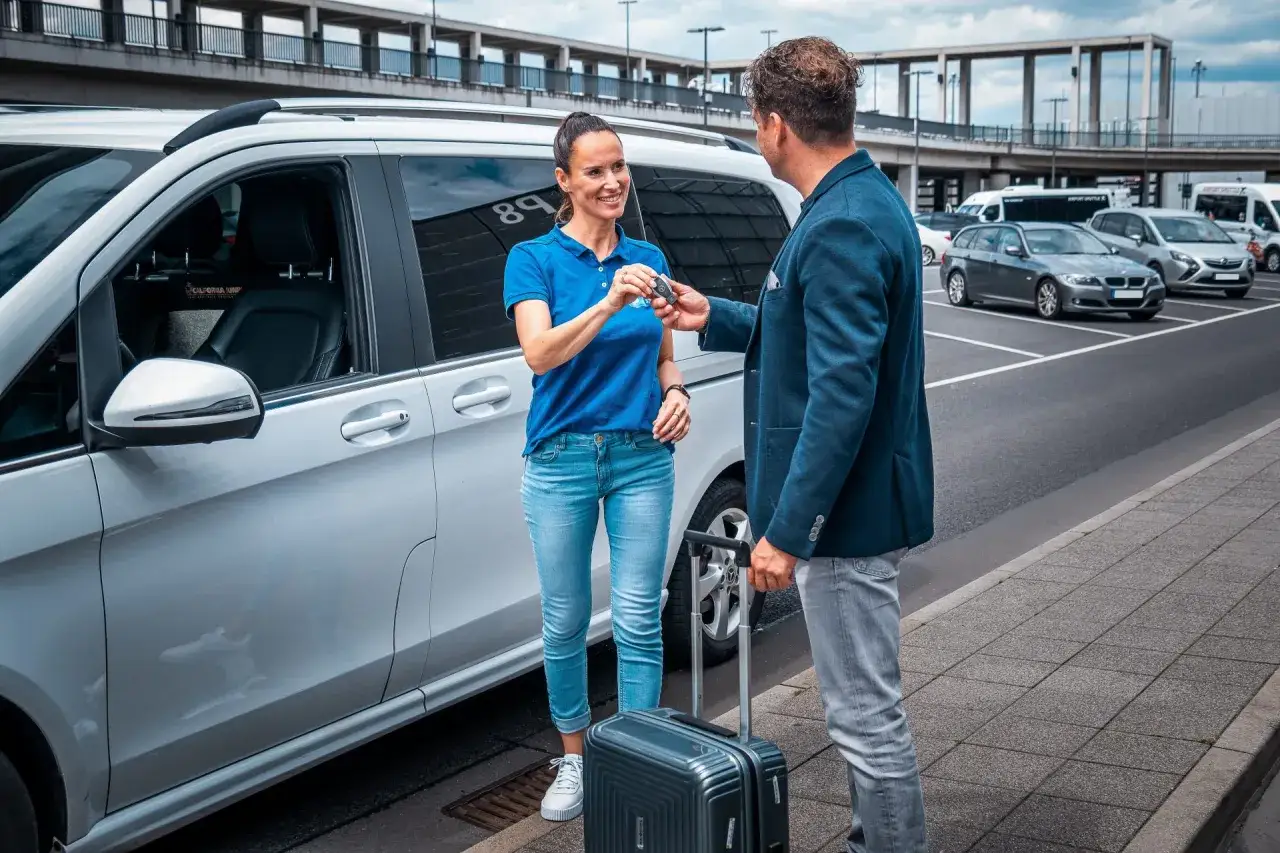 Mann übergibt Schlüssel für Valet Parken an Mitarbeiterin neben weißem Van. Koffer steht bereit.