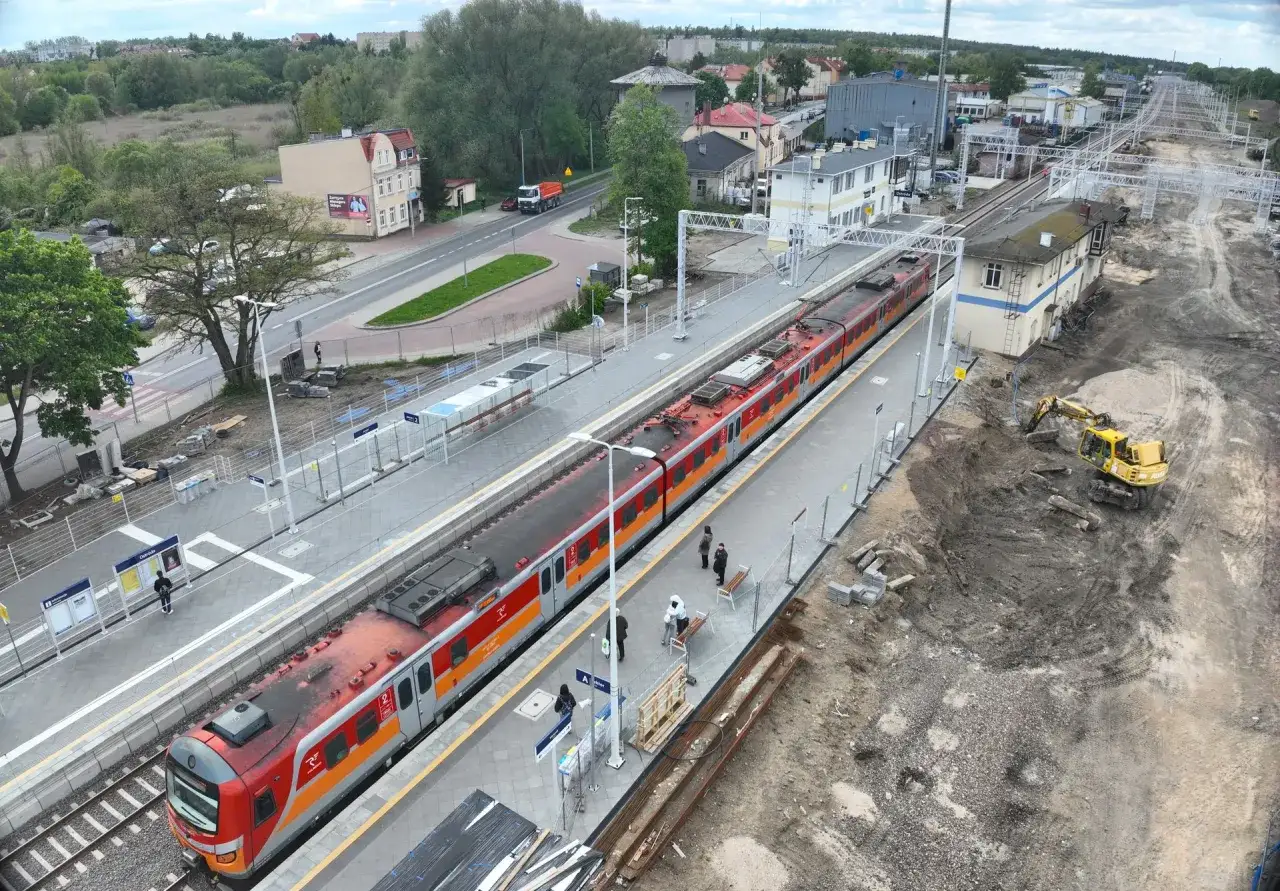 Nowoczesna stacja kolejowa w budowie. Pociąg stoi na peronie, obok koparka i plac budowy.