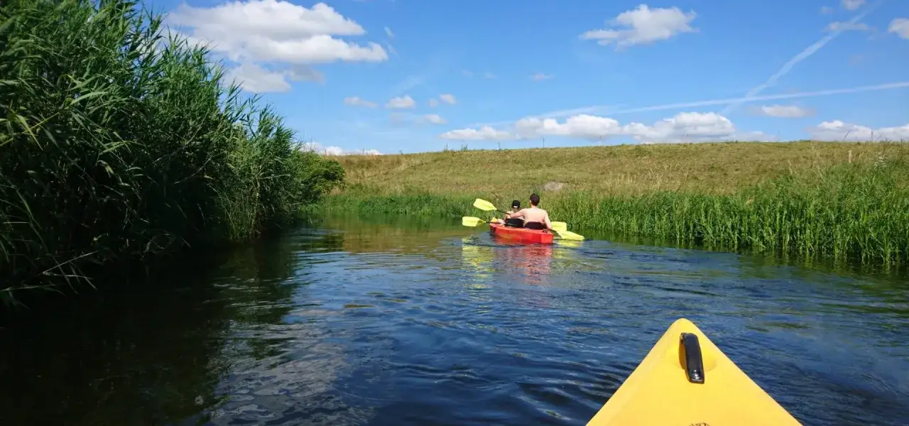 Spływ kajakowy po mazurskich kanałach to wspaniałe mazury atrakcje. Dwie osoby płyną czerwonym kajakiem wśr&oacute;d zielonych trzcin.