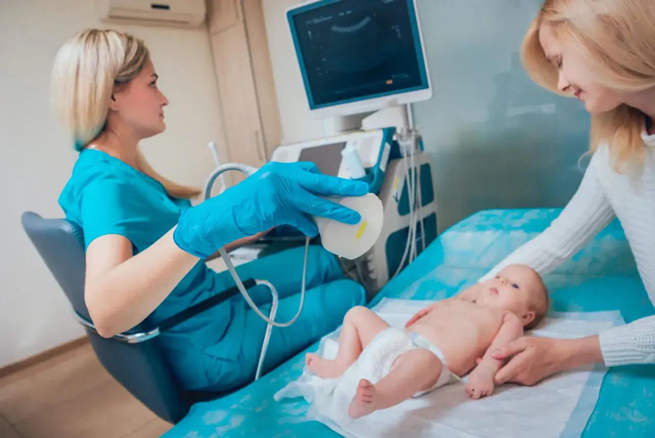 Lekarka bada bioderka u niemowlaka za pomocą aparatu USG. Mama czuwa obok.