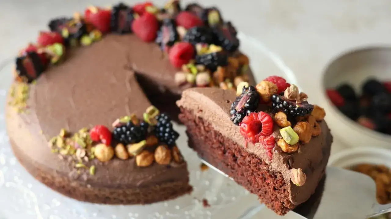 En saftig protein kladdkaka toppad med chokladfrosting, färska bär och nötter. En bit lyfts fram.