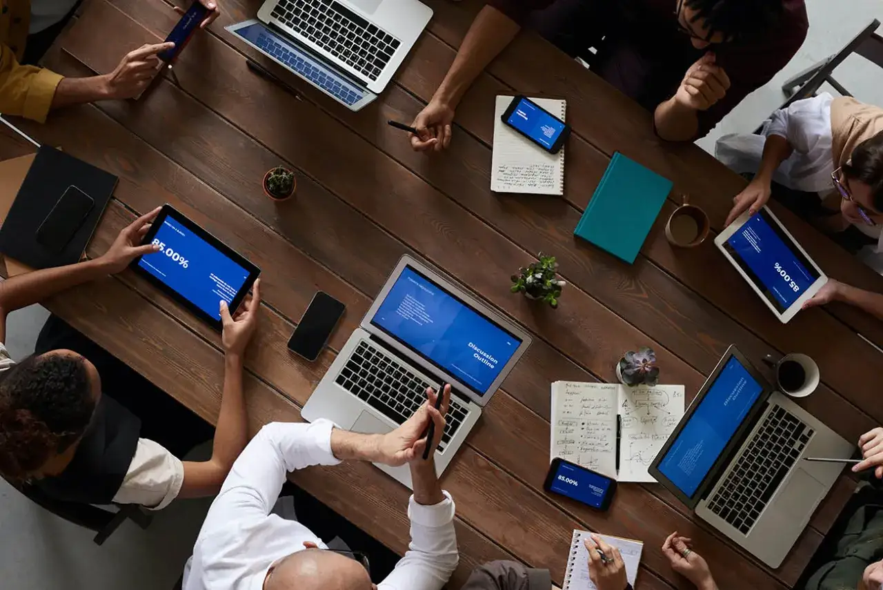 Persone attorno a un tavolo di legno, con laptop, tablet e smartphone. Il **dematerializzazione significato** è evidente: tutto è digitale, niente carta.