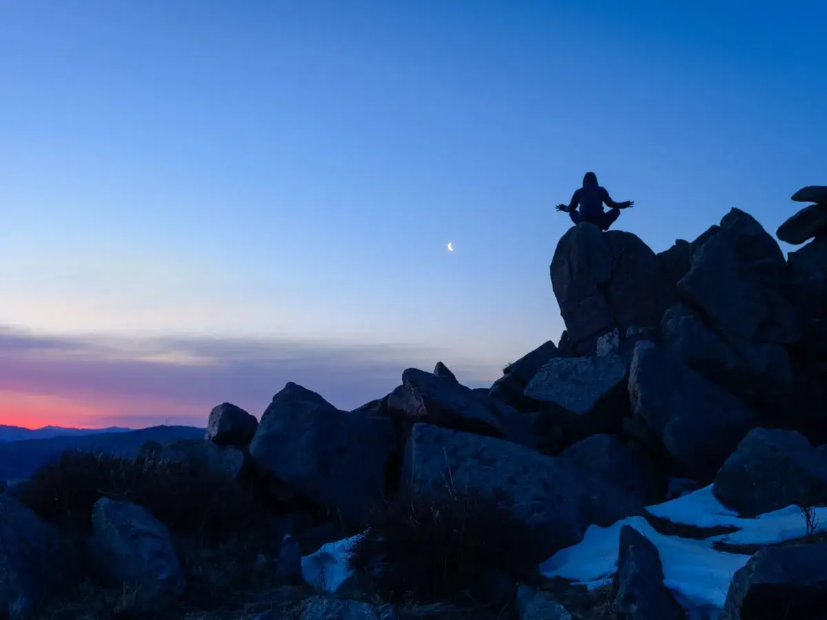 Silueta meditando en la cima de una roca al amanecer, con la luna menguante significado de nuevos comienzos.