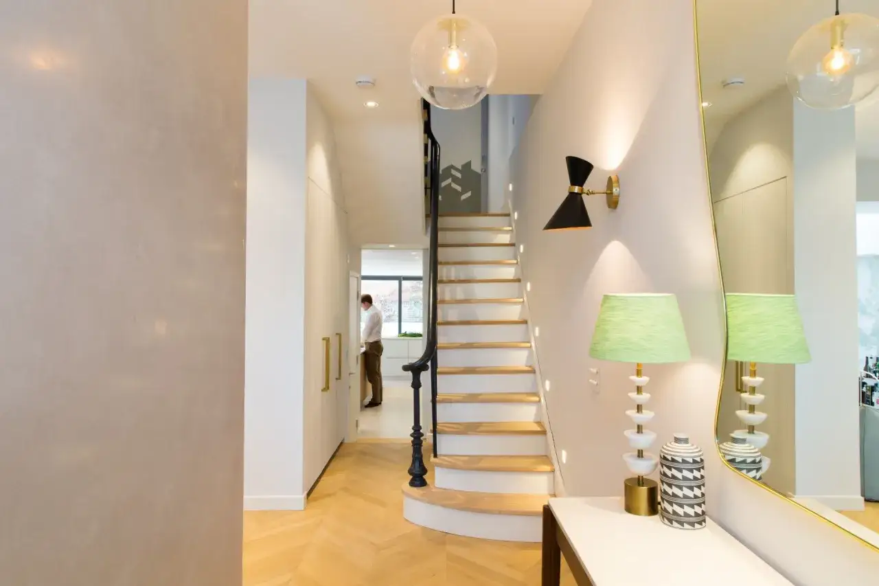 A man stands in a bright kitchen at the end of a **dark hallway**. A modern staircase with wooden steps and a black railing leads up.