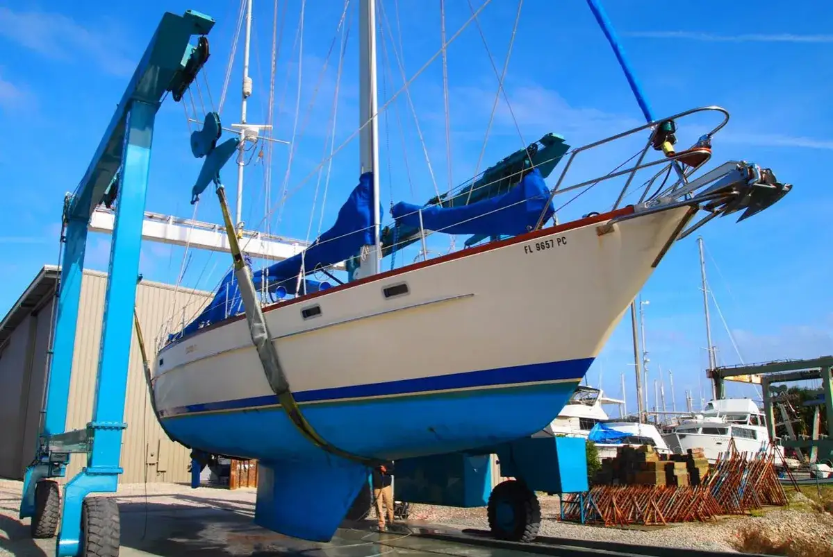 Barca a vela bianca e blu sollevata da una gru blu per una perizia nautica. Ancoraggio a prua, vele blu coperte.