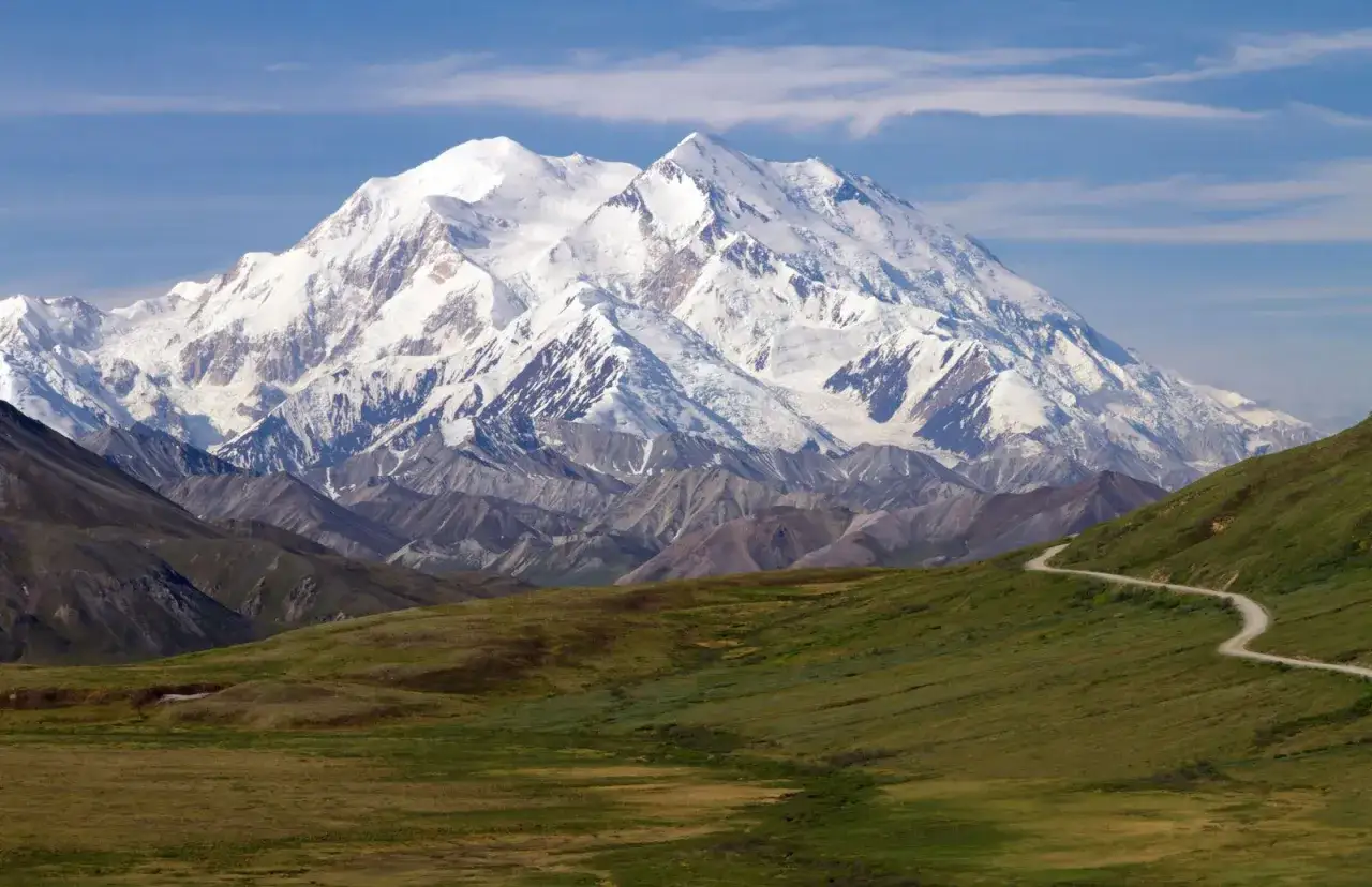 Majestatyczny, ośnieżony najwyższy szczyt Ameryki P&oacute;łnocnej, Denali, g&oacute;ruje nad zielonymi wzg&oacute;rzami i krętą drogą.
