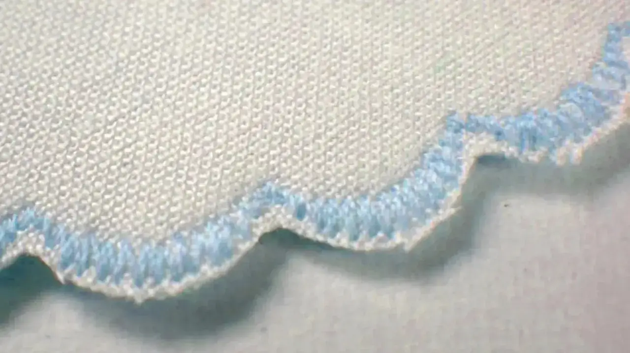 Close-up of a white fabric edge finished with a decorative blue serger stitch, creating a scalloped effect.